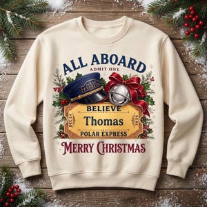 May include: A cream-colored crewneck sweatshirt featuring a graphic print with the text ALL ABOARD ADMIT ONE, BELIEVE, Thomas, POLAR EXPRESS, and MERRY CHRISTMAS. The design includes a navy conductor hat, a silver bell with a red bow, and holly.