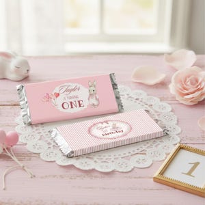 May include: Two pink chocolate bars with silver foil wrapping. One bar features a bunny illustration and text that reads "Taylor is turning ONE." The other bar has a pink striped pattern and text that reads "Taylor's 1st Birthday."