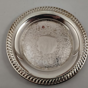 May include: A circular, silver-coloured serving tray with an intricate floral design in the centre. The tray features a raised, braided border and a reflective surface. The tray is likely made of metal.