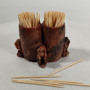 Peut inclure: Un porte-cure-dents décoratif avec deux compartiments cylindriques remplis de cure-dents en bois. Le support est de couleur brun rougeâtre avec un motif marbré et comporte trois petites figurines de singes. Plusieurs cure-dents en vrac sont éparpillés.