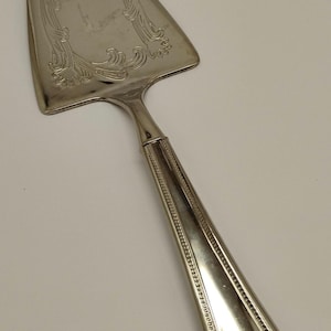 May include: Silver cake server with a decorative floral design on the blade. The handle has a detailed, ridged pattern. A classic serving utensil for cakes and pies.