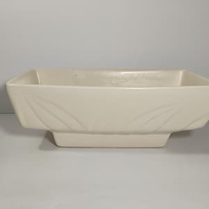 May include: An off-white ceramic planter, rectangular in shape, with a raised base. The sides of the planter are decorated with a delicate leaf pattern. The planter's dimensions make it suitable for a variety of plants or decorative displays.