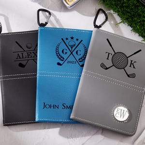 May include: Three personalized golf score card holders in black, blue, and gray. Each holder features a unique design with crossed golf clubs and a golf ball. The black holder has "ALEX" engraved, the blue has "JOHN SMITH" and the gray has initials.