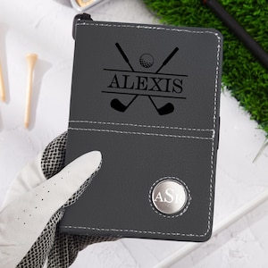 May include: A dark gray golf score card holder with the name "ALEXIS" and golf club graphics. The holder has a silver emblem with the initials "AS". White stitching and a golf glove are visible.