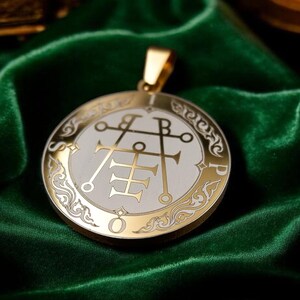 May include: A gold-colored pendant with a white center and intricate symbols. The pendant has a gold bail and is set against a dark green velvet background. The design features a circular shape with ornate detailing.