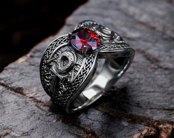 May include: A silver ring featuring a dragon motif and a prominent red gemstone. The ring's design is intricate, with detailed metalwork. The red stone is set in the centre. The ring is displayed on a dark, textured surface.