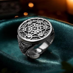 May include: A silver-toned metal ring featuring a circular face engraved with a geometric Metatron's Cube pattern. The band has a polished finish with intricate detailing on the sides, showcasing a complex, symmetrical design in a metallic silver color.