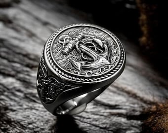 Puede incluir: Un anillo plateado con un diseño de ancla. El anillo presenta un emblema de ancla detallado en el centro, rodeado por un borde en forma de cuerda. El anillo está sobre un fondo oscuro y texturizado, resaltando los intrincados detalles de la joyería de temática náutica.