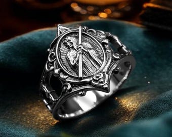 May include: A silver-toned metal ring featuring an oval centerpiece with an embossed figure holding a staff. The band has intricate, openwork scroll detailing along the sides, creating a textured appearance with a polished, reflective finish.