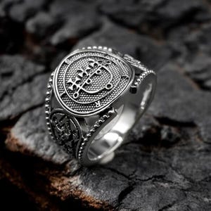 Puede incluir: Un anillo plateado con un diseño intrincado que presenta un emblema central y patrones detallados. El anillo se encuentra sobre un fondo oscuro y texturizado, resaltando su brillo metálico y su artesanía. El diseño del anillo incluye un símbolo central y detalles ornamentados.