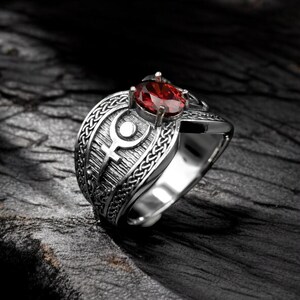 Puede incluir: Anillo plateado con una gran gema roja ovalada. El anillo presenta intrincados diseños grabados en la banda, incluyendo un símbolo central y patrones trenzados. Se encuentra sobre un fondo oscuro y texturizado.