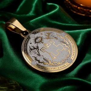 May include: A circular gold-toned pendant featuring an intricate engraved design with a central geometric symbol, an eye motif, and surrounding celestial patterns. The pendant has a polished gold bail at the top and a metallic finish with black detailing.