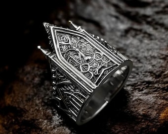 May include: A detailed silver ring with an ornate, gothic cathedral design. The ring features intricate patterns and architectural details, including pointed arches and spires. The ring is set against a dark, textured background.