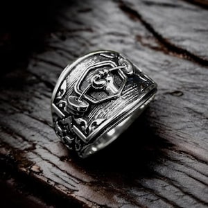 Puede incluir: Un anillo plateado con un diseño detallado que presenta una barra y pesas de halterofilia. El anillo tiene una banda ornamentada y texturizada y está colocado sobre una superficie de madera oscura. El diseño del anillo está centrado en la parte superior.