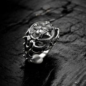 May include: A silver ring with an elaborate design. The ring has a central emblem encircled by flame-like patterns. The ring is set against a dark, textured background, emphasising its intricate details. A gothic-style ring.