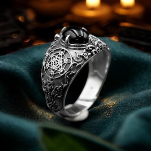 May include: A silver ring featuring a polished black cabochon stone set at the top. The band is decorated with intricate engraved patterns, including a prominent geometric Metatron's Cube symbol on the side, surrounded by detailed scrollwork.