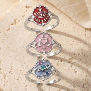 May include: Three silver rings with colorful enamel designs. The rings feature a red crown, a pink heart, and a blue planet. Each ring has a unique design and is set against a neutral background.