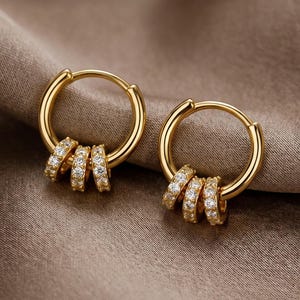 Op de afbeelding: Een paar gouden oorringen. Elke oorring heeft een kleine ring met drie kleinere, met diamanten bezette ringen die eronder hangen. De oorbellen worden gepresenteerd op een bruine, zijdeachtige stof.