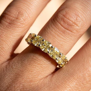 May include: A ring featuring a continuous band of cushion-cut yellow stones set in a silver-toned metal. The stones are arranged side-by-side in a prong setting, creating a uniform row of bright yellow gems that encircle the finger completely.