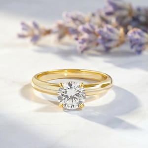 May include: A gold ring featuring a single round brilliant-cut clear gemstone set in a four-prong setting. The band is smooth and polished, showcasing a classic circular design that highlights the reflective facets of the central stone.
