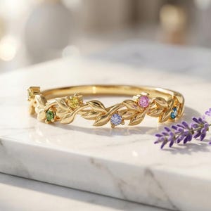 Puede incluir: Una pulsera dorada con un diseño de hojas y piedras preciosas. La pulsera presenta pequeñas piedras preciosas de colores verde, amarillo, rosa, azul y turquesa. Las hojas están intrincadamente detalladas. La pulsera se exhibe sobre una superficie de mármol.