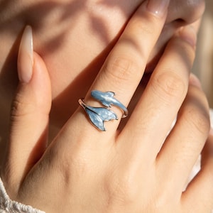 May include: A silver-toned open ring featuring a light blue enamel design shaped like a dolphin. The ring wraps around the finger, with the dolphin's head and tail forming the two ends of the band, showcasing a smooth, glossy finish.