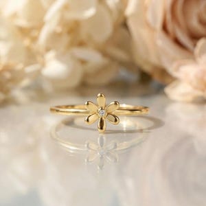Puede incluir: Un anillo dorado con un diseño de flor de margarita y una pequeña gema transparente en el centro. El anillo se encuentra sobre un fondo floral suave y borroso. La banda del anillo es fina y delicada.