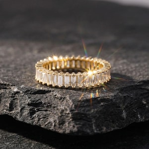 Op de afbeelding: Een gouden eternity ring met rechthoekig geslepen, heldere edelstenen. De ring ligt op een donkere, getextureerde ondergrond. De edelstenen zijn dicht rond de band geplaatst.