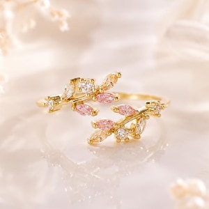 May include: A gold-toned open ring featuring two branch-like ends adorned with marquise-cut pink stones and small round clear stones. The design mimics a delicate vine, with the metal band curving to create an adjustable, open-ended silhouette.