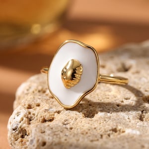 Op de afbeelding: Een goudkleurige ring met een ovaal ontwerp van wit emaille en een getextureerd gouden middenstuk. De ring is geplaatst op een getextureerd stenen oppervlak, wat het unieke ontwerp benadrukt. De ringband is ook goudkleurig.