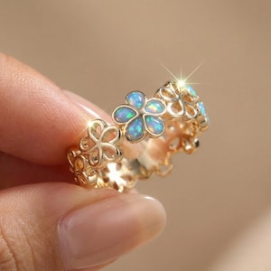 May include: A gold-colored ring featuring a floral design. Each flower has a blue opal center. The ring is in focus, with the details of the flowers and the gold band clearly visible. The ring is a piece of jewelry.