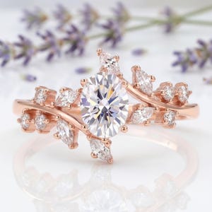 Pode incluir: Um anel em ouro rosa com uma pedra preciosa central transparente de corte oval. A banda apresenta um design de ramo, adornado com várias pedras transparentes mais pequenas de corte marquise e redondo ao longo da pedra central.