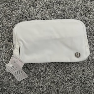 May include: A white rectangular belt bag with a zip closure and a small silver logo. The bag has a tag attached with text. The bag is made of a smooth, possibly nylon, material.