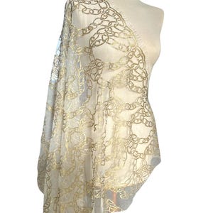 Può includere: Un tessuto trasparente bianco sporco drappeggiato su un manichino, con un intricato motivo a catena dorata. Il materiale delicato ha un bordo sfrangiato ed è ideale per creare abiti eleganti o accenti decorativi.