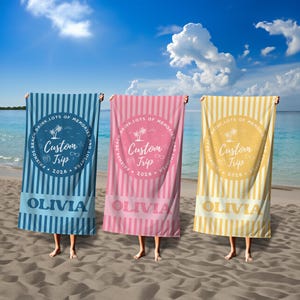 May include: Three beach towels in blue, pink, and yellow, each with vertical stripes. The towels feature a circular design with text that reads "Custom Trip 2026" and the name "Olivia". The background is a beach scene.