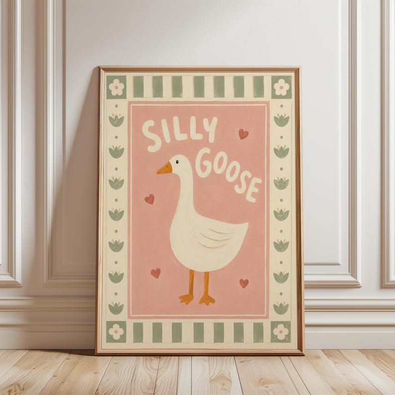 May include: A framed wall art print featuring a white goose with an orange beak and feet on a dusty pink background. The text "SILLY GOOSE" is written in white above the goose, surrounded by small red hearts. A border of green stripes and flowers.