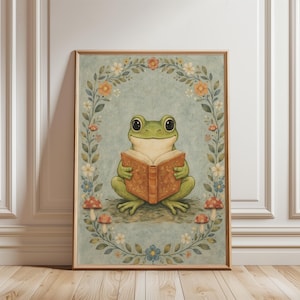 May include: A framed art print featuring a whimsical illustration of a green frog reading a book. The frog is surrounded by a floral wreath with mushrooms at the base. The background is a soft blue-green, and the frame is a light wood color.