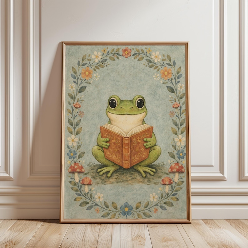May include: A framed art print featuring a whimsical illustration of a green frog reading a book. The frog is surrounded by a floral wreath with mushrooms at the base. The background is a soft blue-green, and the frame is a light wood color.