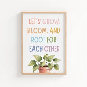 May include: A framed print with the words "LET'S GROW, BLOOM, AND ROOT FOR EACH OTHER" in pastel colors. A watercolor illustration of a potted plant with green leaves and a terracotta pot is at the bottom of the print. The frame is a light wood color.