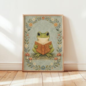 May include: A framed art print featuring a green frog reading a book. The frog is surrounded by a floral wreath with mushrooms at the bottom. The background is a light blue color, and the frame is a light brown color.