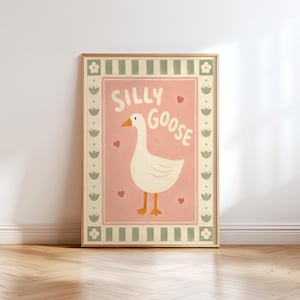 May include: A framed wall art print featuring a white goose with an orange beak and feet centered on a dusty pink background. The text SILLY GOOSE is printed in white above the goose, surrounded by small red hearts and a sage green floral border.