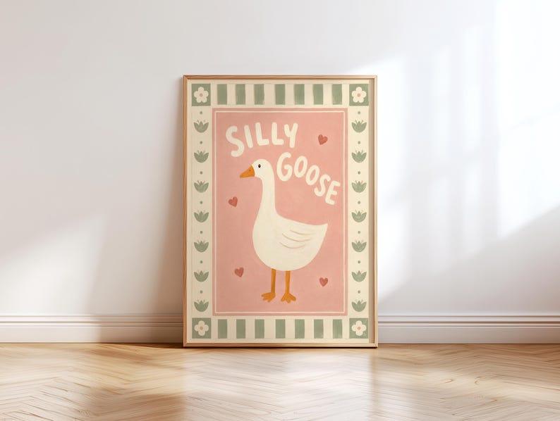May include: A framed wall art print featuring a white goose with an orange beak and feet centered on a dusty pink background. The text SILLY GOOSE is printed in white above the goose, surrounded by small red hearts and a sage green floral border.