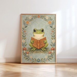 May include: A framed art print featuring a whimsical illustration of a green frog reading a book. The frog is surrounded by a floral wreath with mushrooms at the base. The artwork has a vintage, storybook aesthetic, with soft colors and a light blue background.