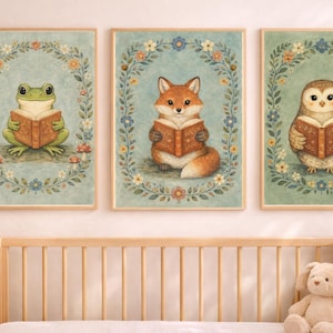 May include: Three framed art prints featuring whimsical animal illustrations. A green frog, a fox, and an owl are each depicted reading a book. Each animal is surrounded by a floral wreath on a light blue background. The prints are displayed above a wooden crib.