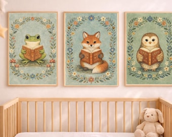 May include: Three framed art prints featuring whimsical animal illustrations. A green frog, a fox, and an owl are each depicted reading a book. Each animal is surrounded by a floral wreath on a light blue background. The prints are displayed above a wooden crib.