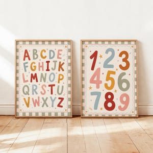 May include: Two framed educational prints. One displays the alphabet in colorful letters, the other shows numbers 1-9. Both have a cream background with a green and white striped border and star accents. The frames are a light brown.