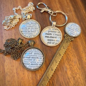 Pode incluir: Uma coleção de joias e acessórios, incluindo colares, um porta-chaves e um marcador de livros. Os pingentes apresentam texto de um dicionário. O marcador de livros é uma régua com um laço decorativo. Os itens são em tons de ouro e prata.