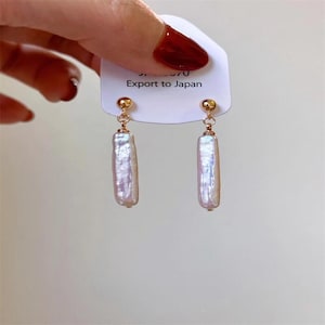 Op de afbeelding: Een paar goudkleurige oorhangers met rechthoekige, iriserende, lichtpaarse parels. De oorbellen hangen aan kleine goudkleurige bolvormige studs. De tekst "Export to Japan" is gedrukt op een wit label.