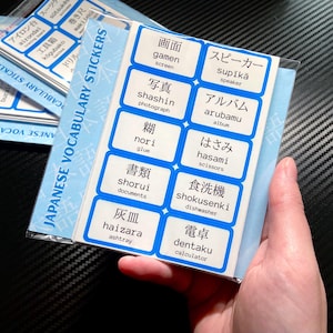 May include: A pack of Japanese vocabulary stickers featuring white rectangular labels with blue borders. Each sticker displays a Japanese kanji character, its romaji transliteration, and the English translation for common household items.