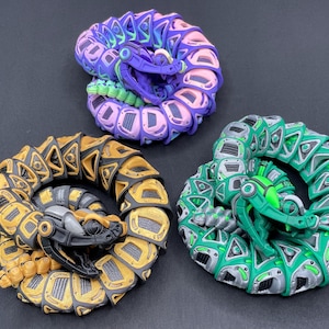 May include: Three coiled, articulated snake sculptures in vibrant colours. One is gold and black, another is green and grey, and the third is purple, pink, and blue. Each has a detailed, mechanical design.